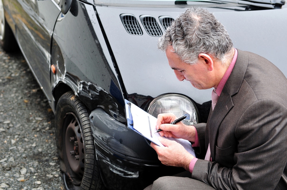 Insurance-representative-writing-on-clipboard-while-looking-at-damaged-car-after-collision