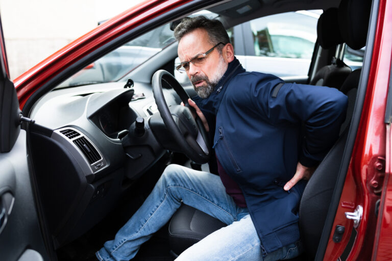 Man with pained expression sitting in driver's seat, holding back