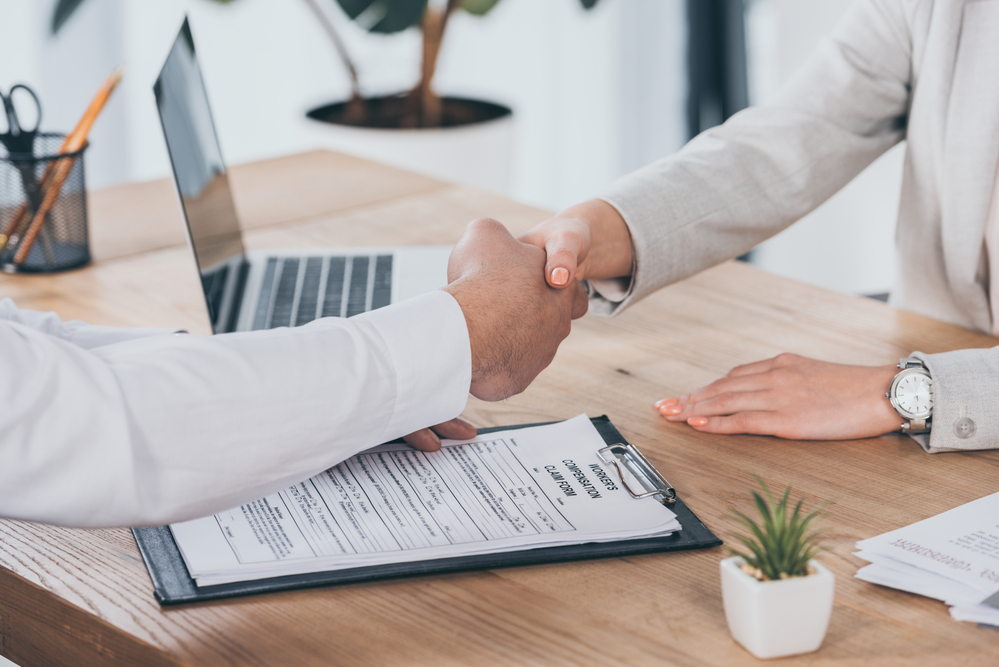 Two professionals shaking hands over desk, workers compensation form on desk