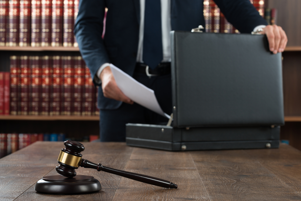 lawyer putting documents in briefcase, gavel on desk, books behind