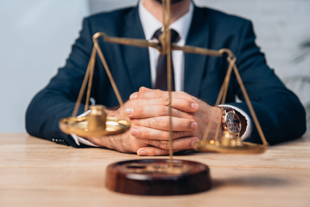 cropped attorney with hands clasped on desk, behind scales of justice