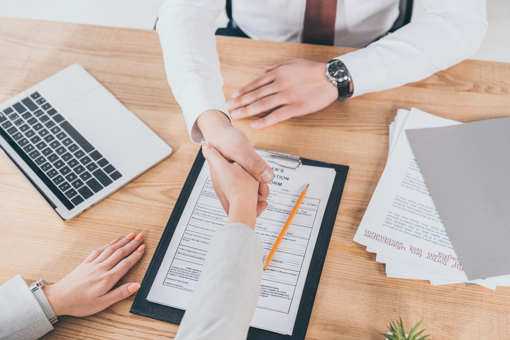 Two people in business attire shaking hands above desk with workers compensation form