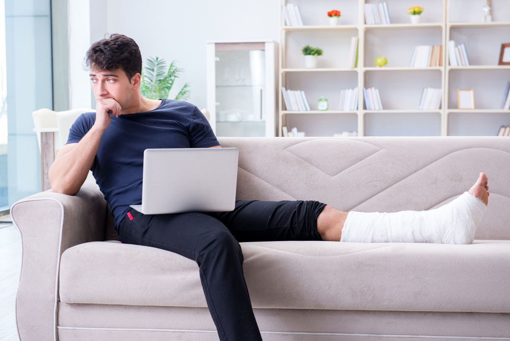 Young man sitting on couch with laptop, his leg in cast