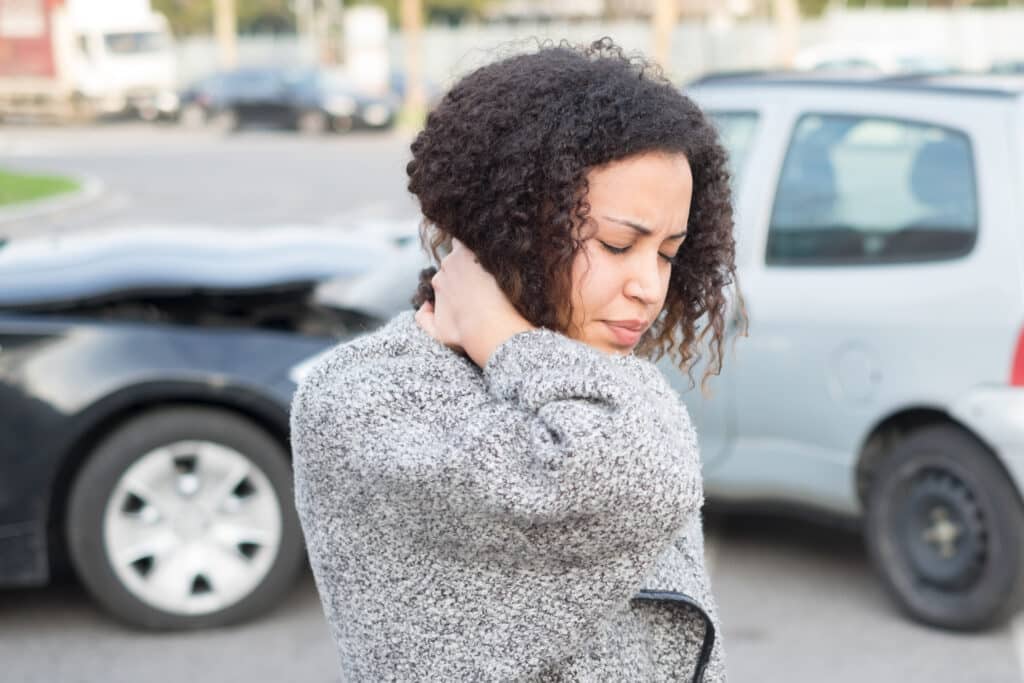 Young woman holding back of neck in pain, car crash in background