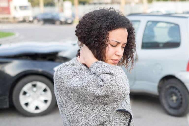 Young woman holding back of neck in pain, car crash in background