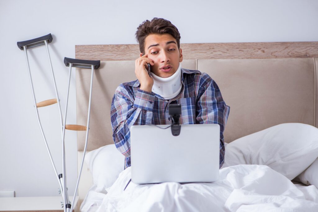 Injured man talking on phone in bed, crutches beside him, wearing neck brace