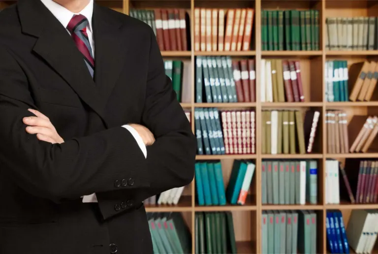 Torso of man in suit arms folded in front of bookcase